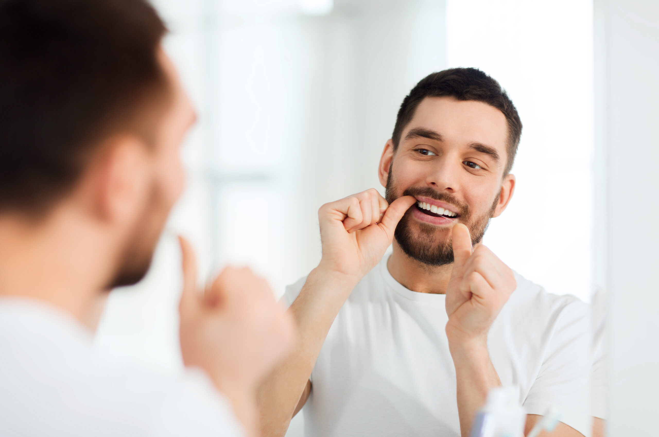 health care, dental hygiene, people and beauty concept - smiling young man with floss cleaning teeth and looking to mirror at home bathroom