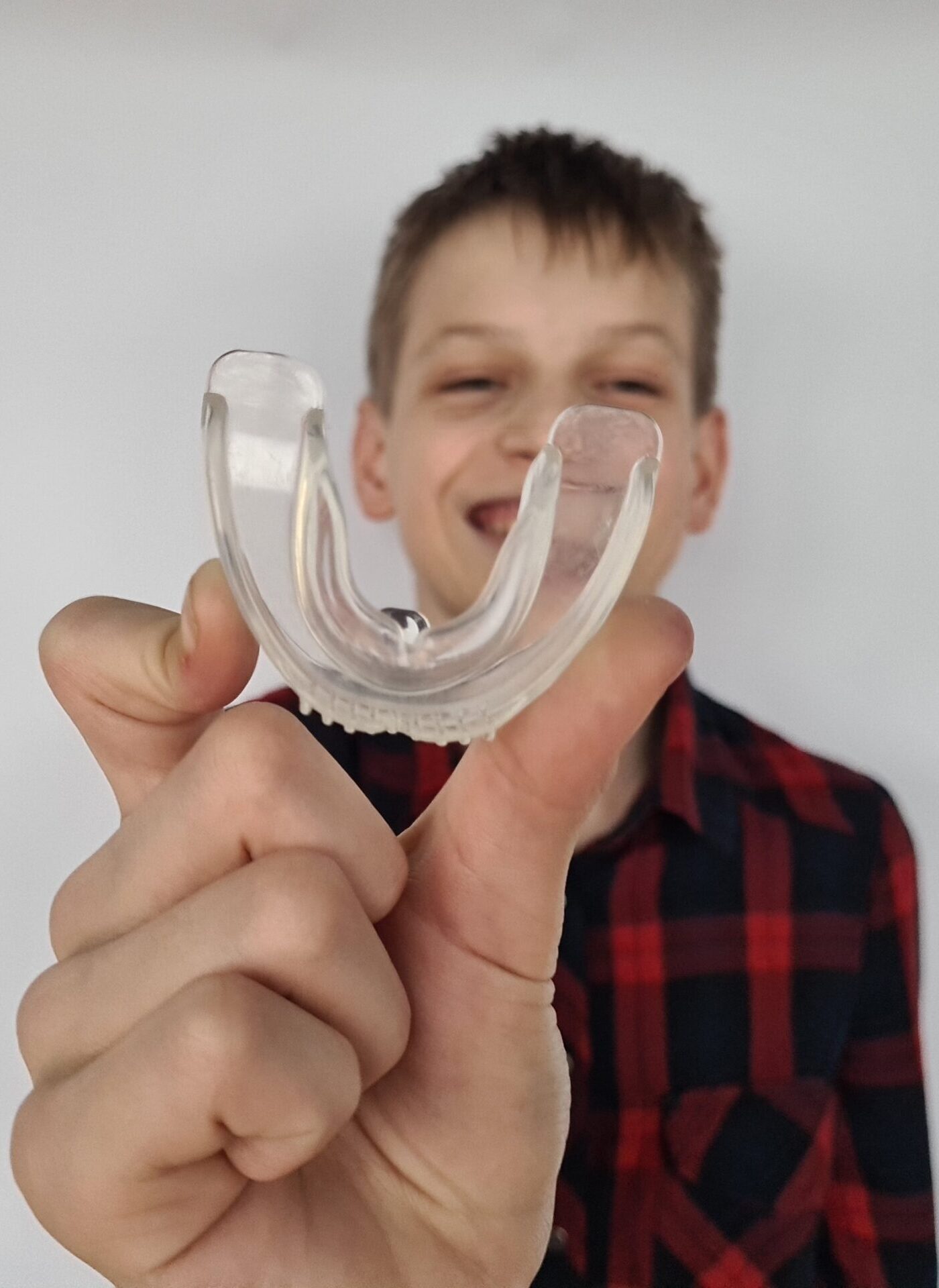 A boy smiles while holding a clear mouthguard in front of a plain background, illustrating dental care and sports safety