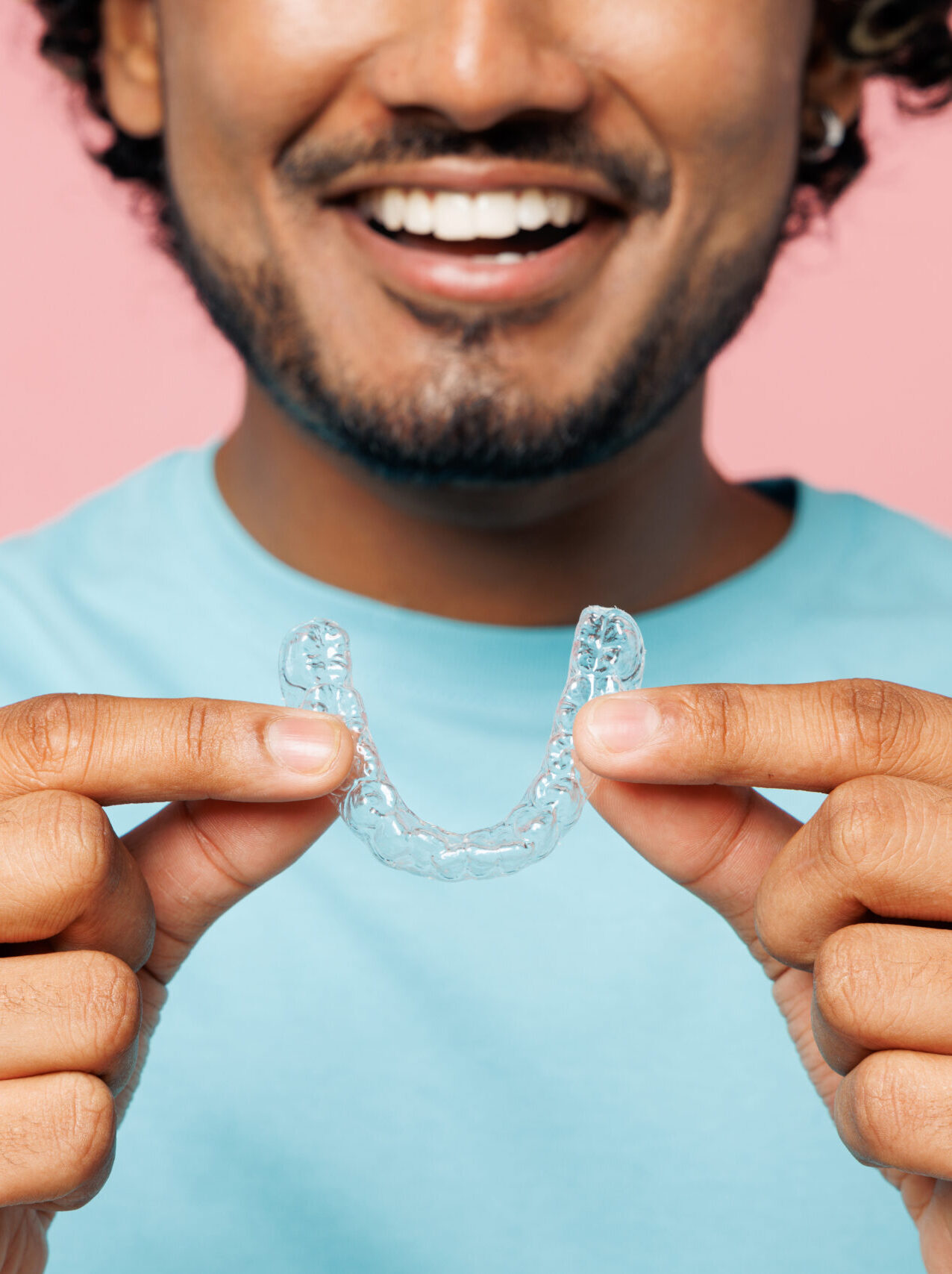 Cropped young smiling Indian man he wear blue t-shirt casual clothes hold in hand invisible transparent aligners, invisalign dental braces isolated on plain pastel pink background. Lifestyle concept