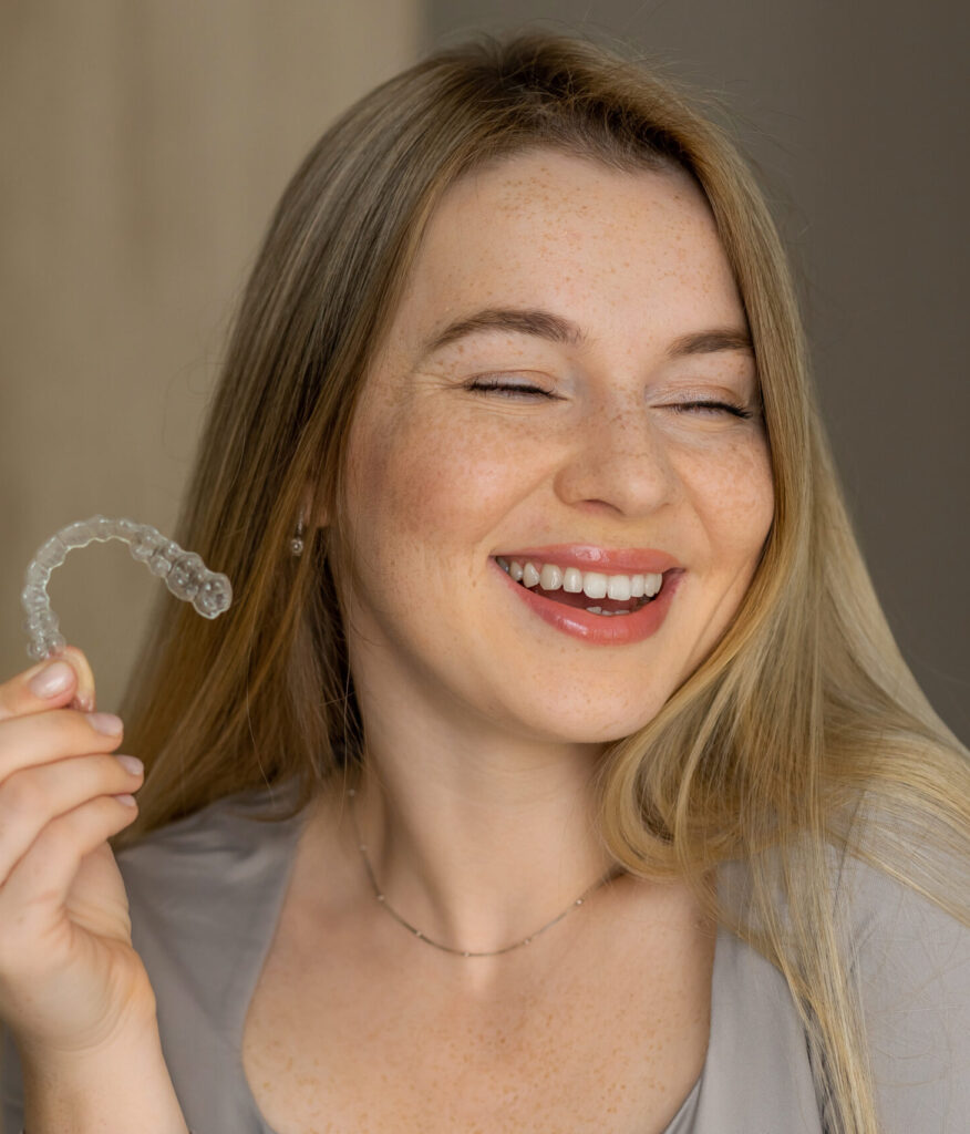 A cheerful young woman is smiling while holding a dental aligner for orthodontic treatment.