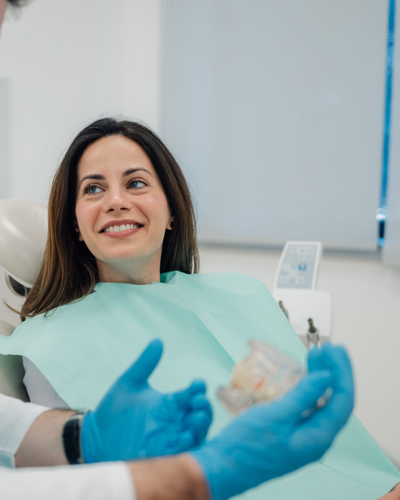 Dentist wearing blue gloves showing dentures to a smiling patient sitting in a dentist chair in a dental clinic, discussing a treatment plan for dental health and oral hygiene
