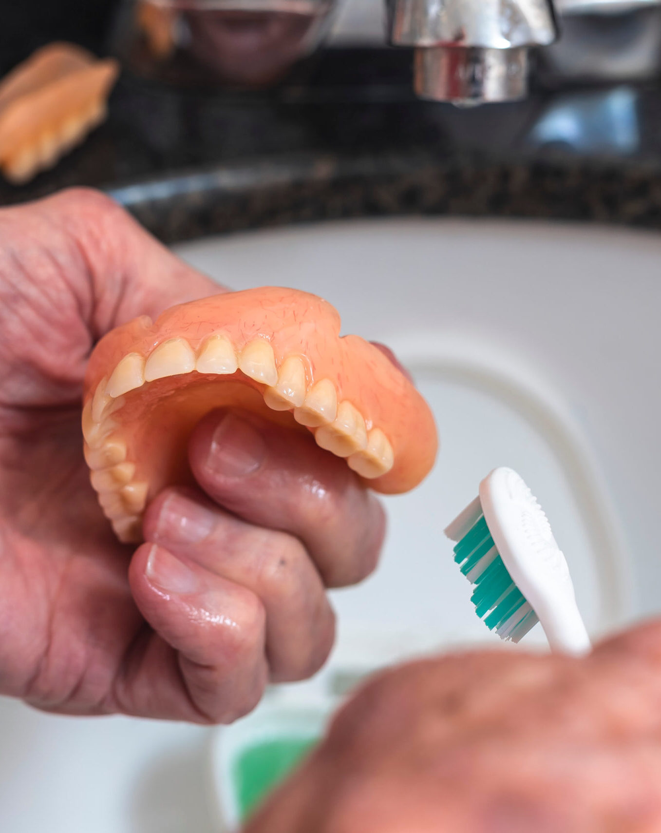 An elderly person holds a full upper denture while cleaning it with a toothbrush over a denture soaking case filled with green cleaning solution, illustrating proper hygiene care.