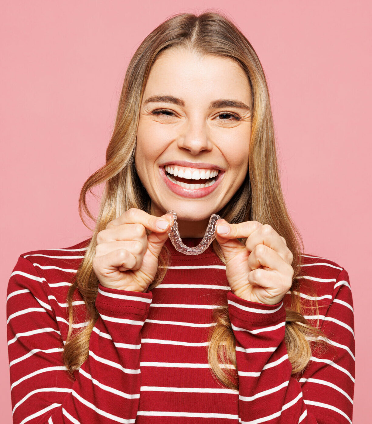 Young smiling satisfied woman wearing red striped sweatshirt casual clothes hold in hand invisible transparent aligners, invisalign dental braces isolated on plain pink background. Lifestyle concept