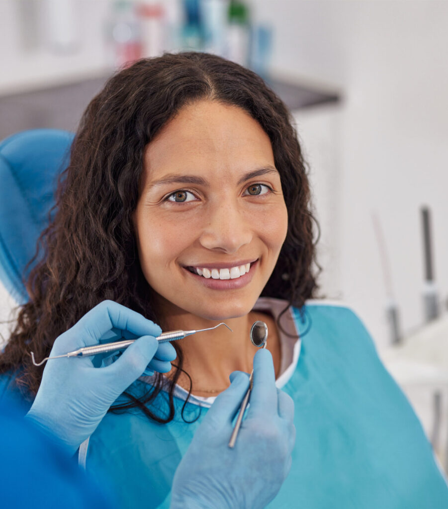 Happy young patient smiling while sitting in dental chair during oral hygiene. Young woman with curly hair wearing dental bib in bright clinic. Dentist holding tools near patient during consultation for whitening teeth treatment.