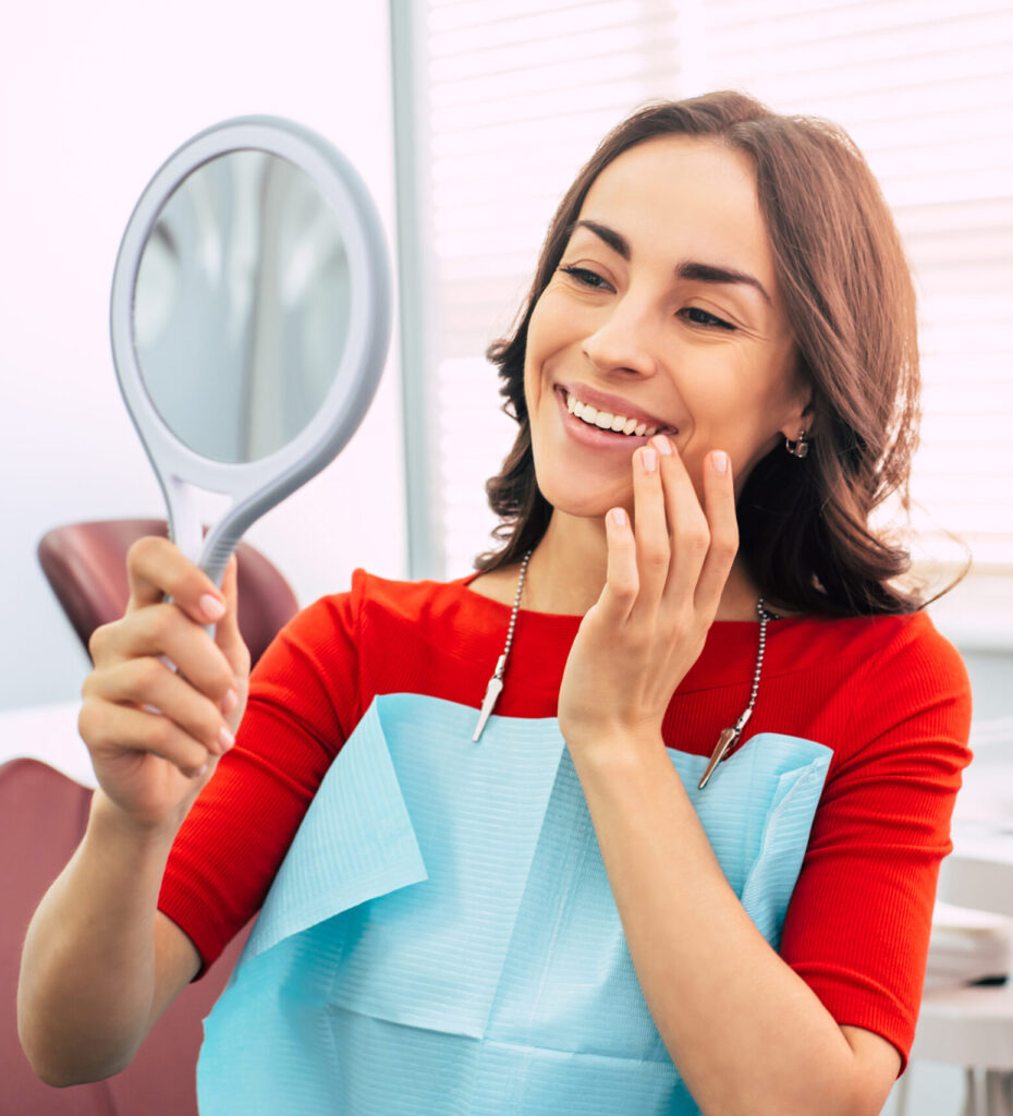 Brilliant work. Pretty girl in dentist cabinet is using a mirror to look through the ideal work of a doctor. She has a special dentist napkin over her vibrant red sweater.