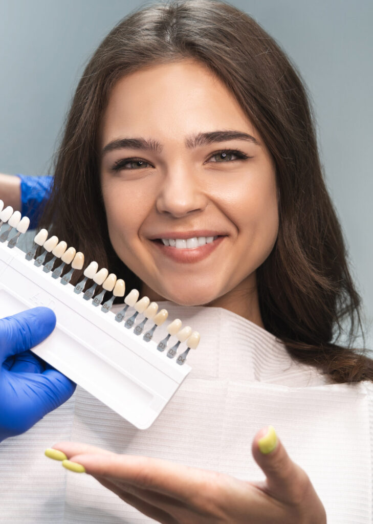 smiling brunette woman patient having appiontment in dental clinic picking up shade using tooth enamel scale held by dentist in blue gloves.