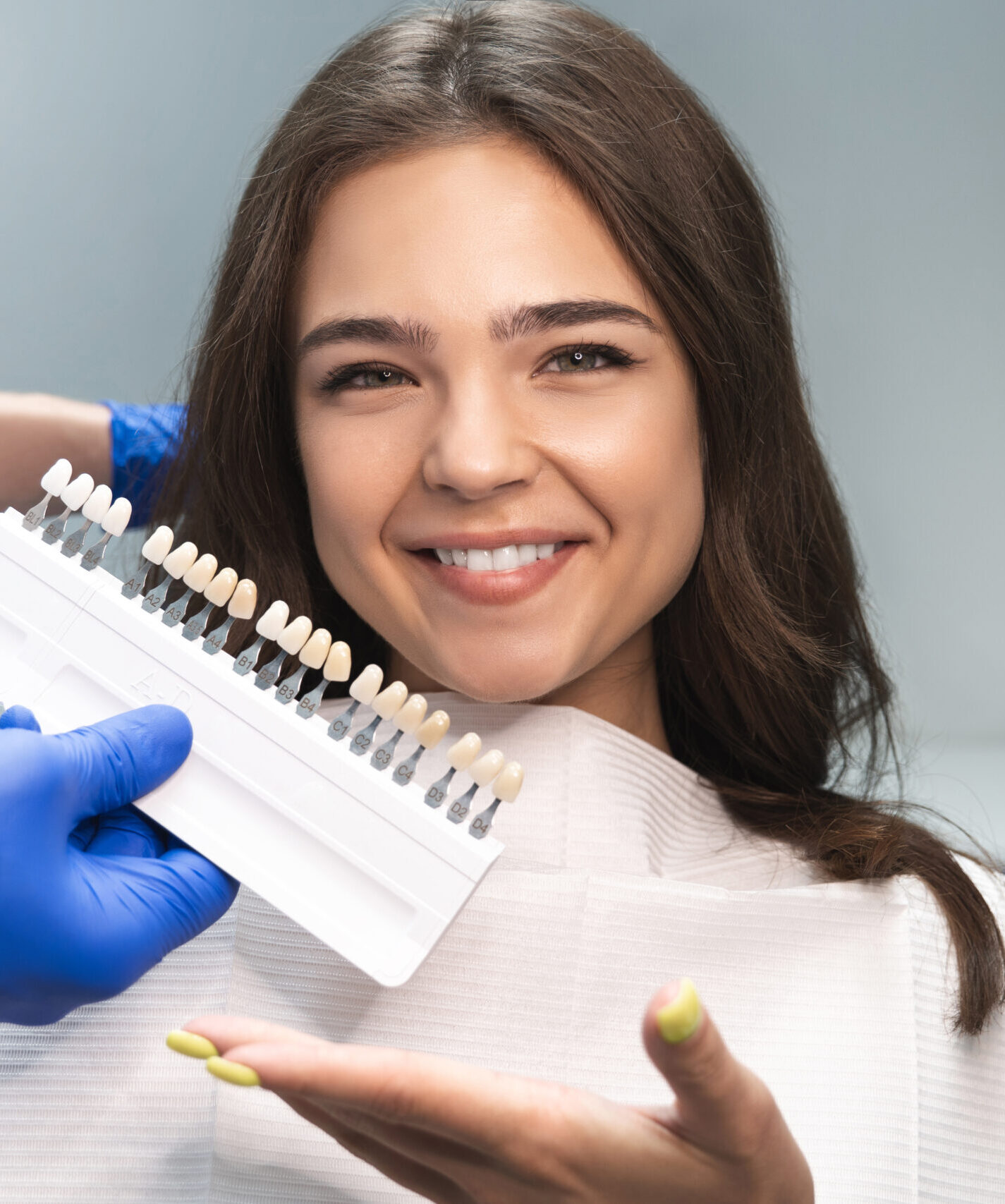 smiling brunette woman patient having appiontment in dental clinic picking up shade using tooth enamel scale held by dentist in blue gloves.
