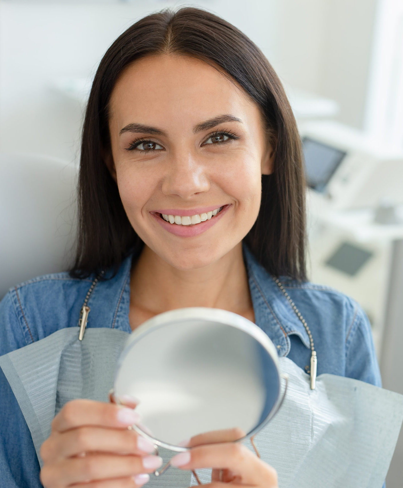 smiling woman sitting in dental chair holding mirror after dental procedure and looking at the camera