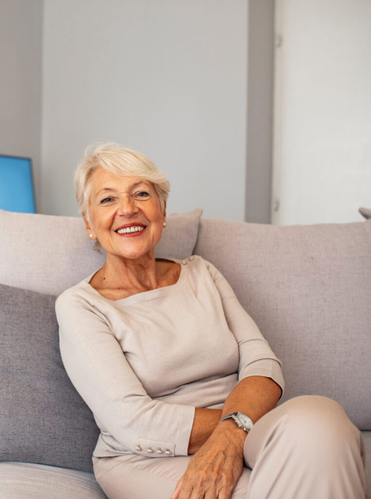 Portrait of beautiful senior woman with white hair. Portrait of senior woman sitting on sofa at home. Smiling middle aged mature grey haired woman looking at camera