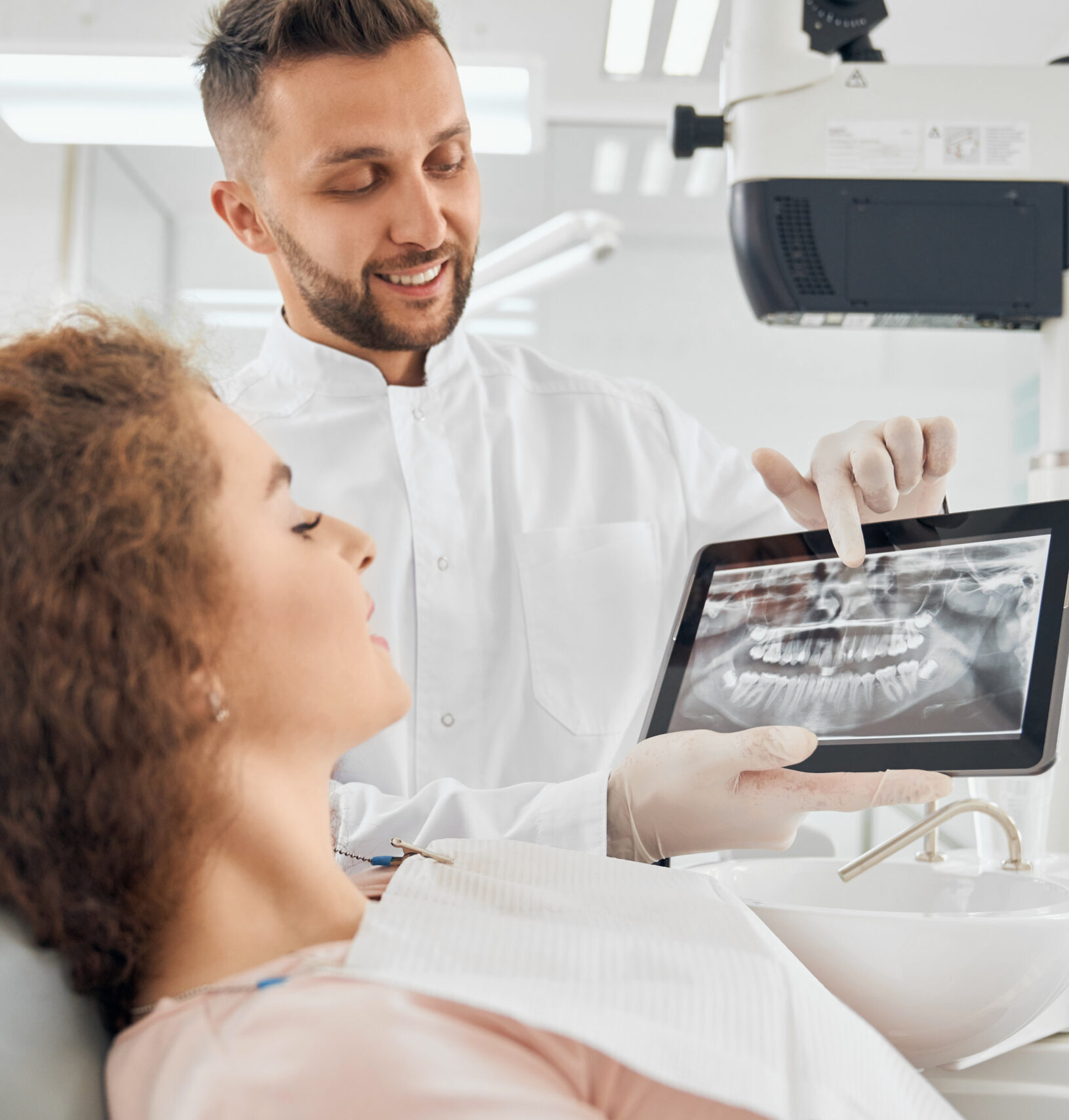 Side view of beautiful girl with curly hair sitting in dental chair and looking at electronic device that holding handsome male doctor. Professional dentist showing x-ray pictures of teeth to client