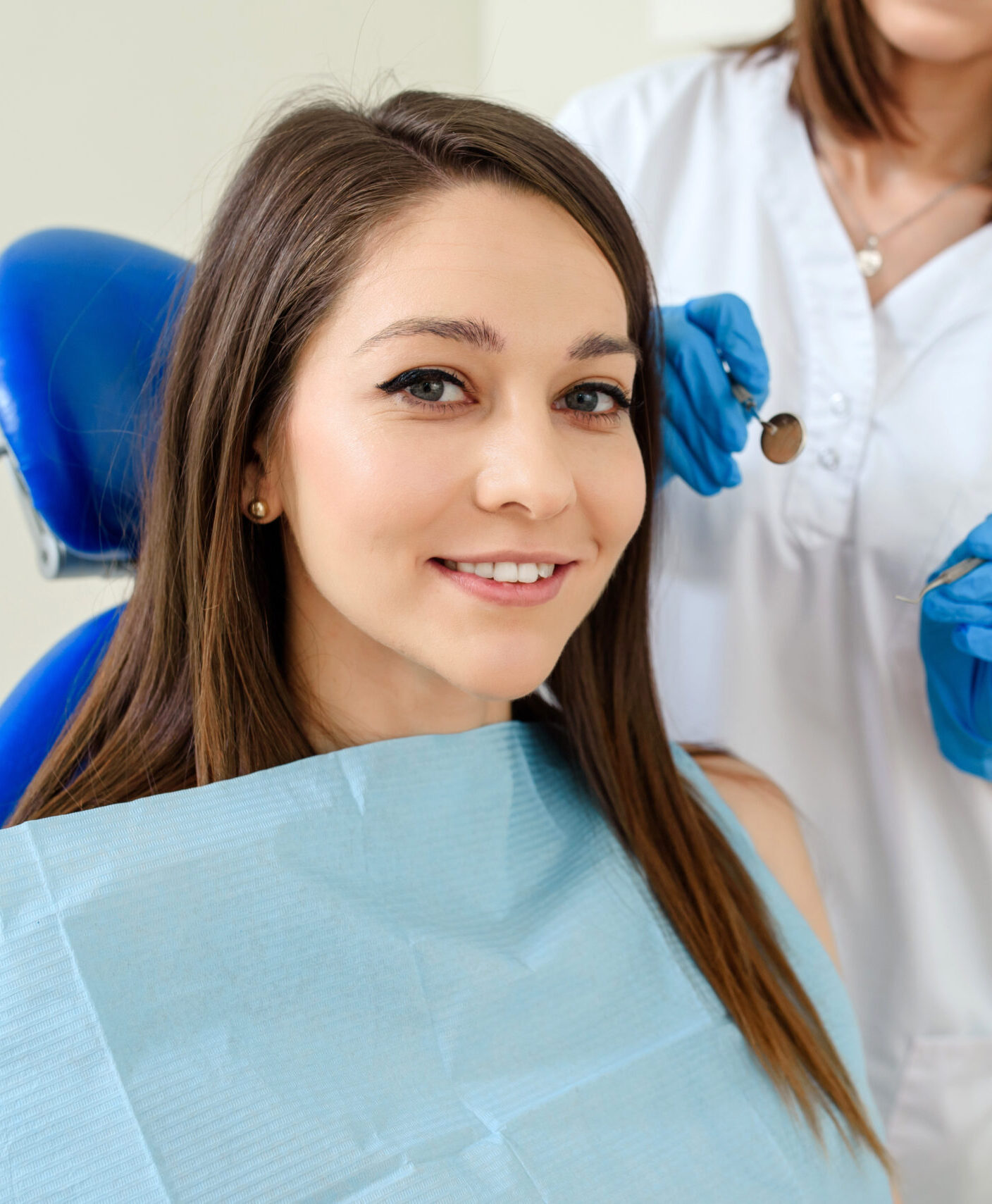 Young happy smiling woman patient in a dental clinic is receiving a dental treatment and looking at the camera. The dentist with dental equipment are behind