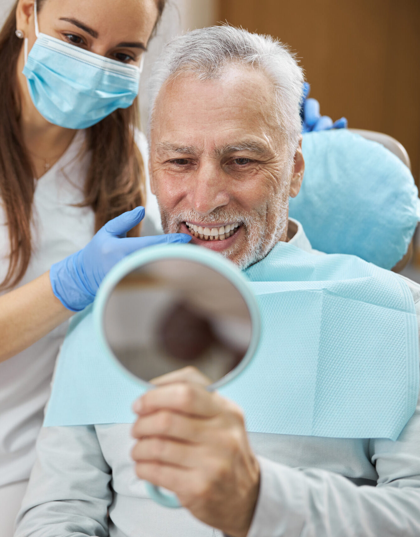 Elderly man and his young female dentist smiling while looking in the mirror in dental clinic