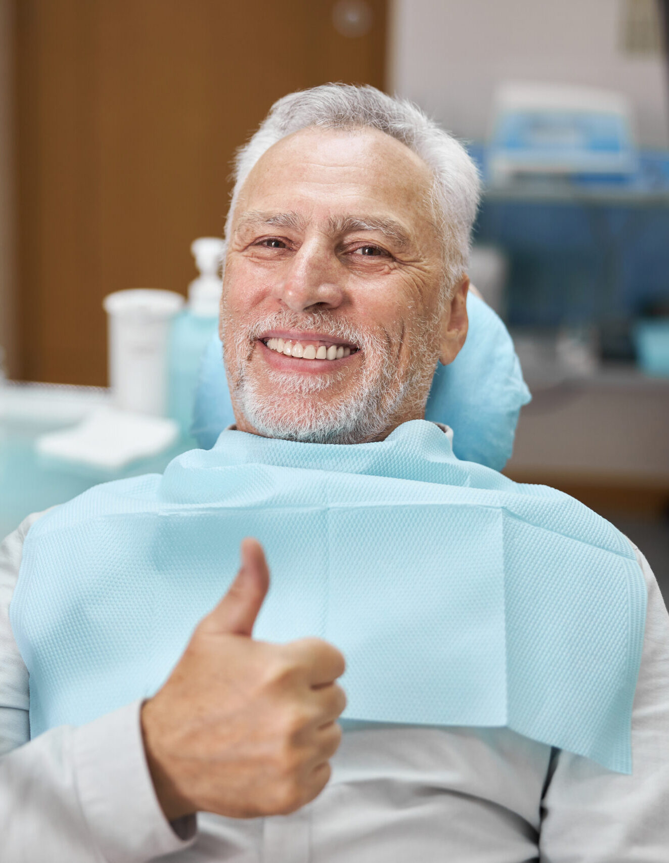 Mirthful senior citizen smiling and showing thumbs up at dental appointment with his dentist and looking at the camera