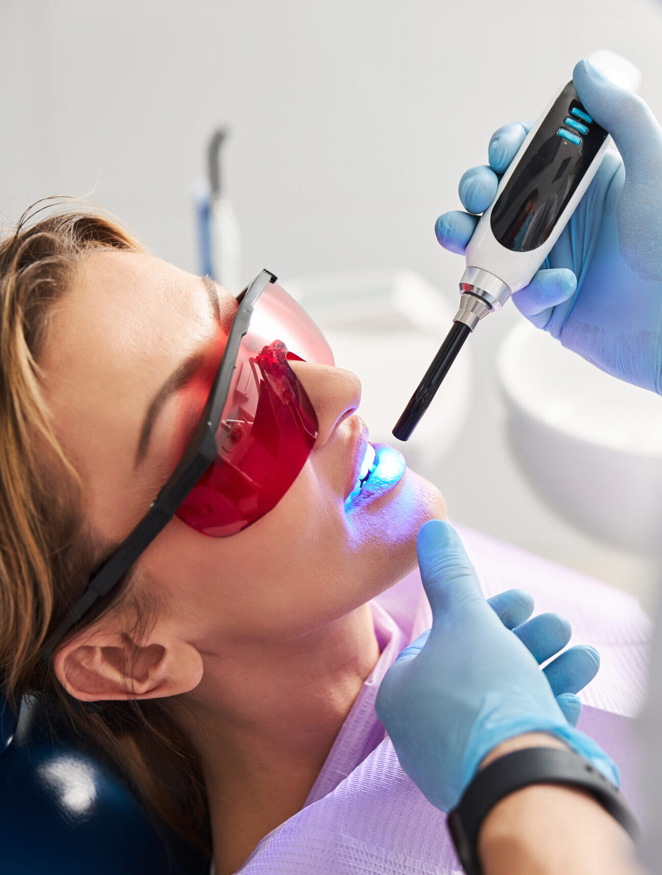Medic shining with LED curing light on lady teeth for hardening of dental fillings while woman wearing red safety glasses