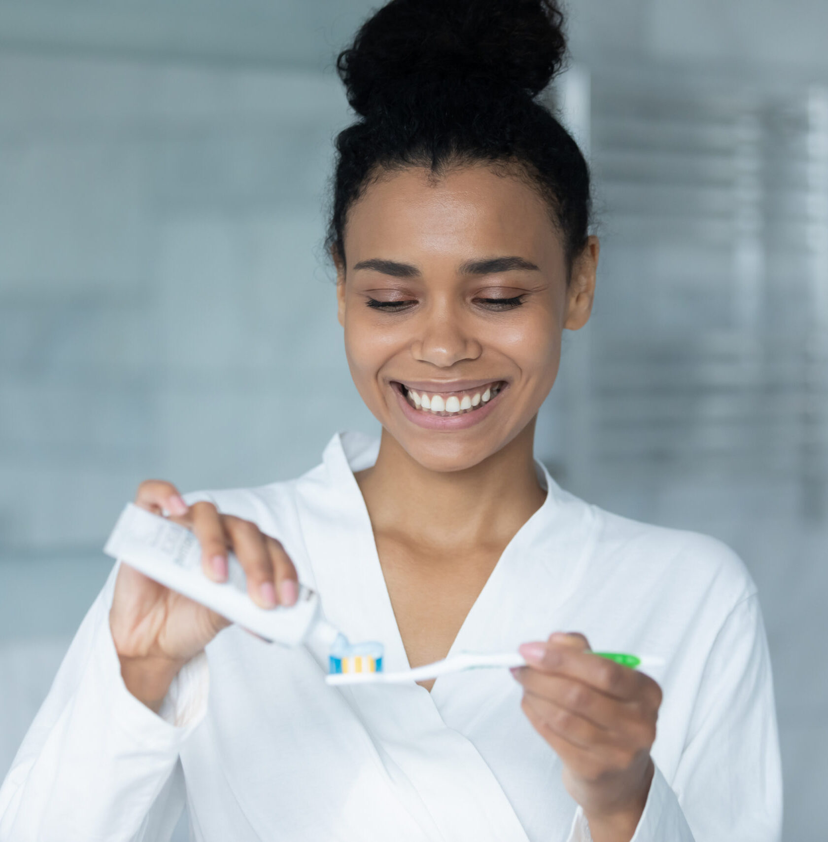 Happy young African American woman squeezing mint toothpaste from tube on plastic toothbrush for brushing teeth in bathroom, keeping morning routine for dental care, healthy enamel, mouth hygiene