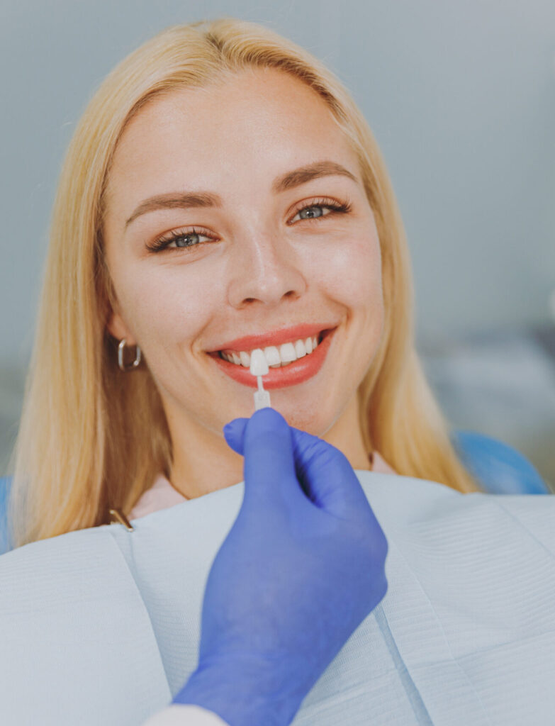 Young smiling calm happy woman 20s choose veneers enamel color compare teeth shade with stomatologist look at result mirror sit at dentist office chair indoor modern cabinet Enamel whitening treatment