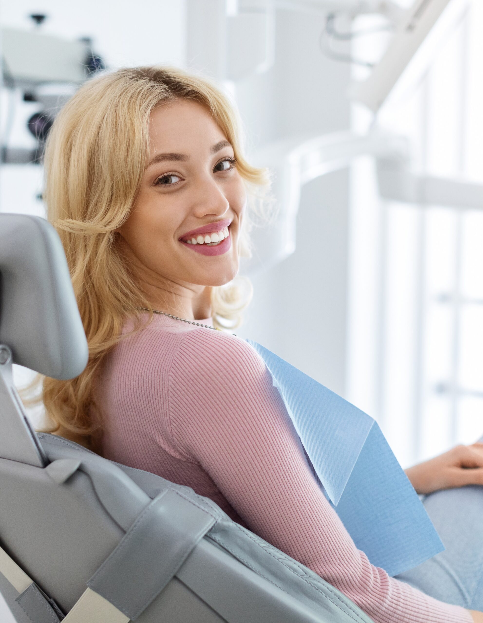 Smiling and relaxed young woman sitting at dental chair in modern clinic, waiting for her dentist. Happy attractive blonde lady attending luxury dental clinic, bacl view, copy space