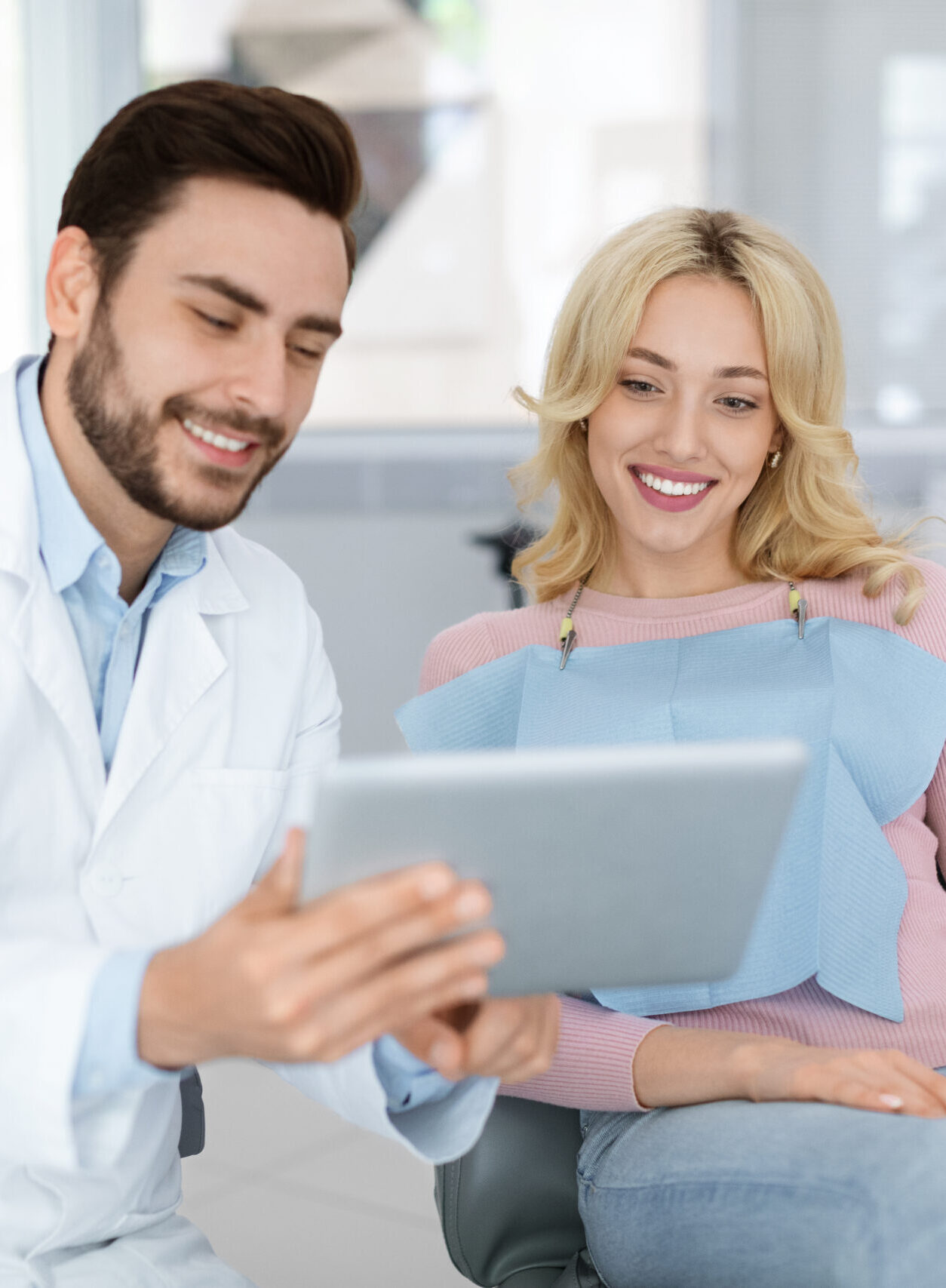 Cheerful young bearded man dentist discussing treatment method with beautiful blonde lady patient sitting at dental chair, looking at digital tablet. Modern technologies and healthcare concept