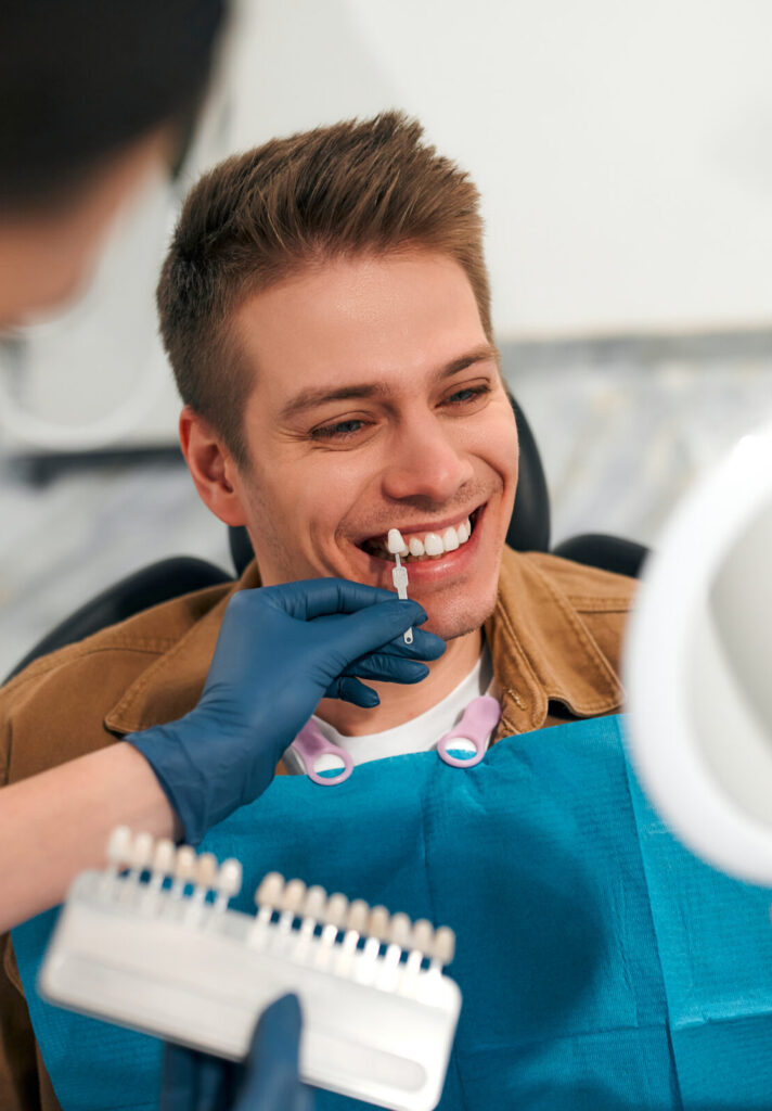 Medicine, dentistry and healthcare concept - closeup of a dentist with tooth color samples choosing a shade for a male patient's teeth in a dental clinic looking at a mirror.