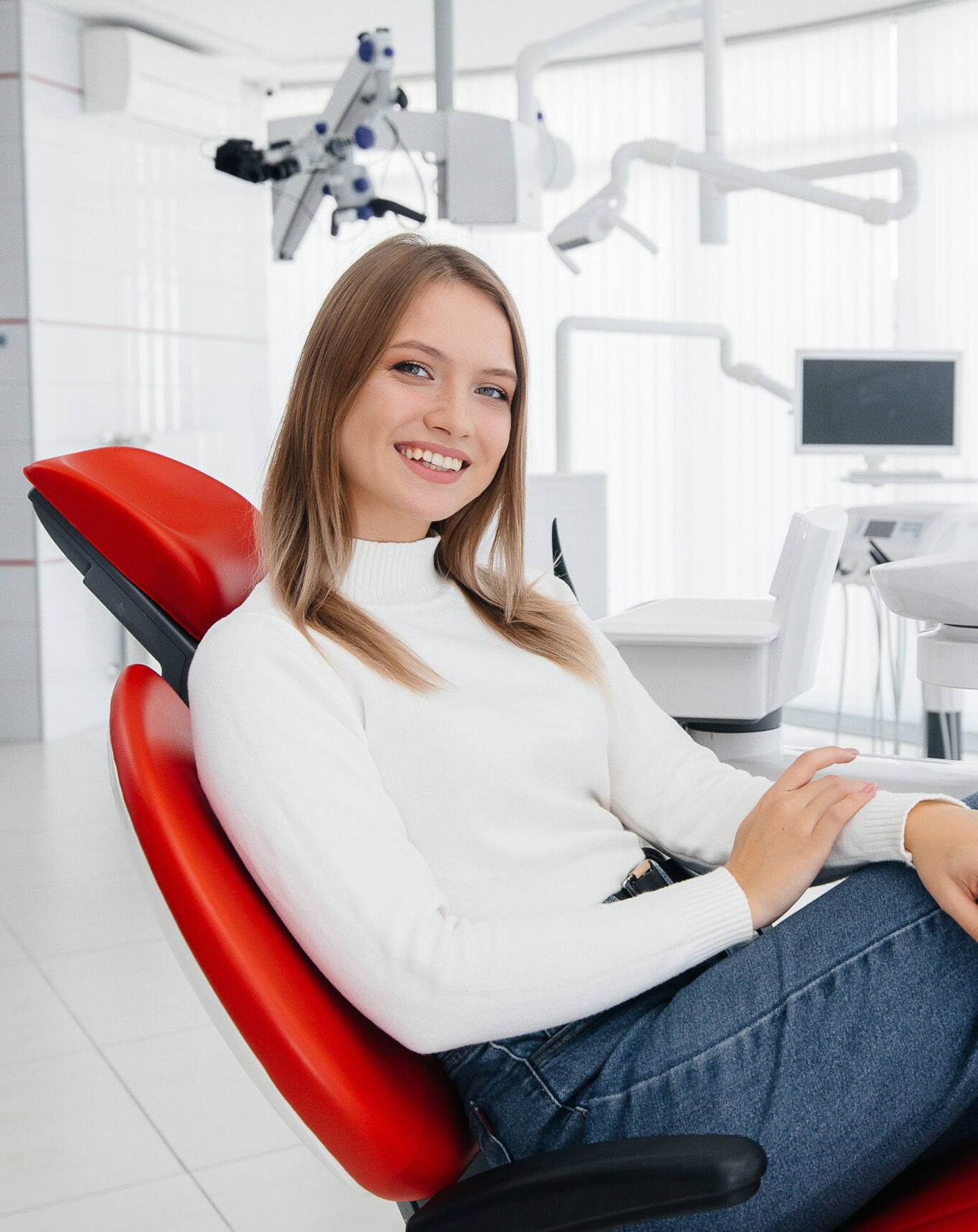 A beautiful young woman is sitting on a red dental chair in modern white dentistry and smiling. Dental prosthetics, treatment and teeth whitening.