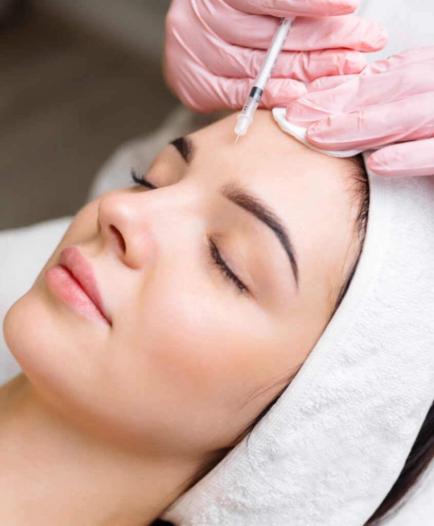 Close up of hands of young cosmetologist injecting botox in female face. She is standing and smiling. The woman is closed her eyes with relaxation.