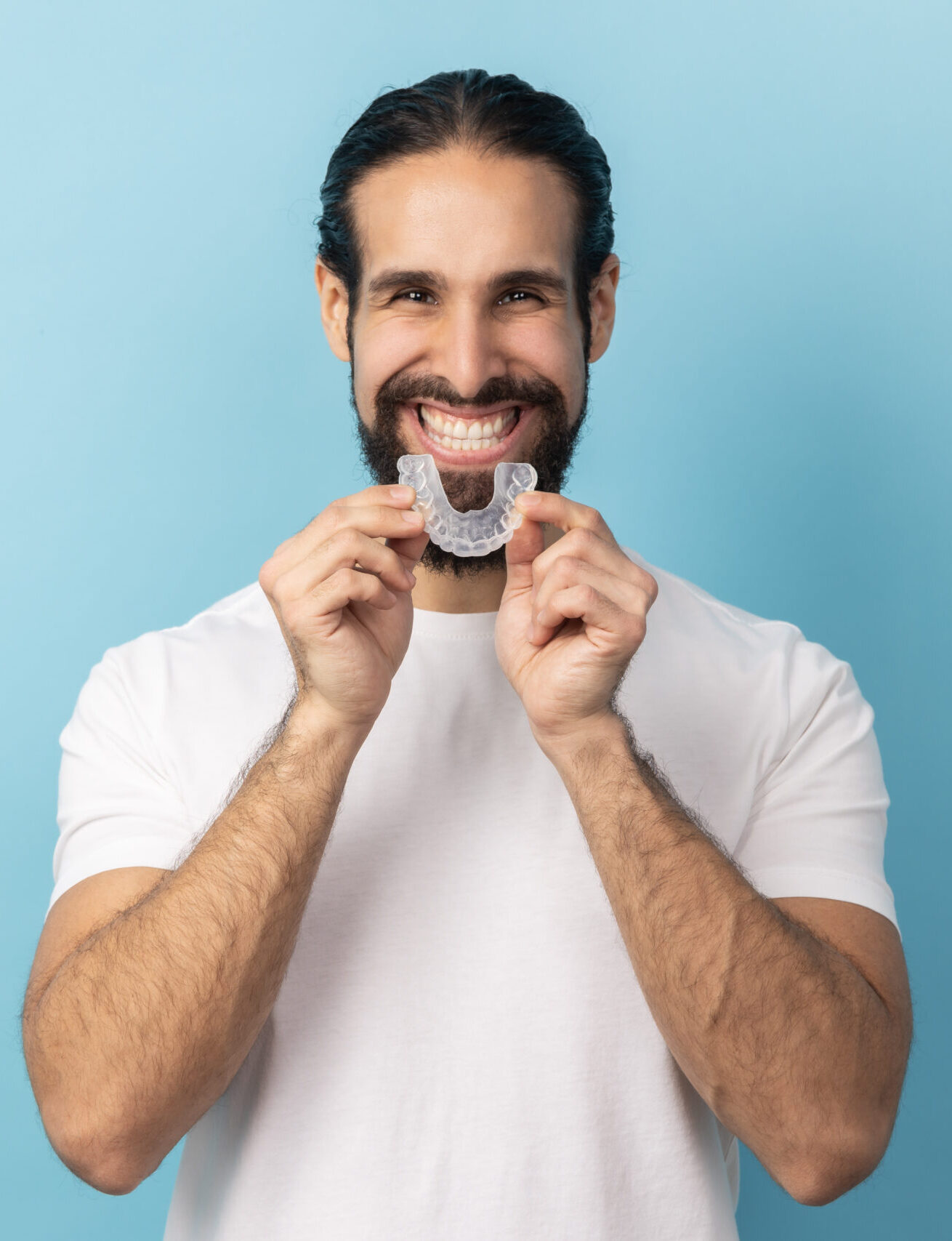 Portrait of man with beard in white T-shirt using teeth whitening braces, dental aligner retainer, dental clinic for beautiful teeth, treatment course. Indoor studio shot isolated on blue background.