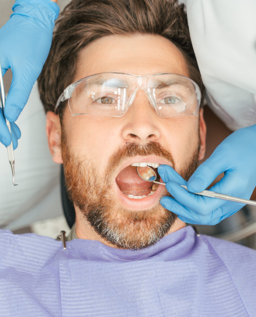 Portrait of handsome bearded man patient sitting in dental chair wearing protective glasses with open mouth, closeup. Female dentist treats teeth with instruments. Concept of dental care