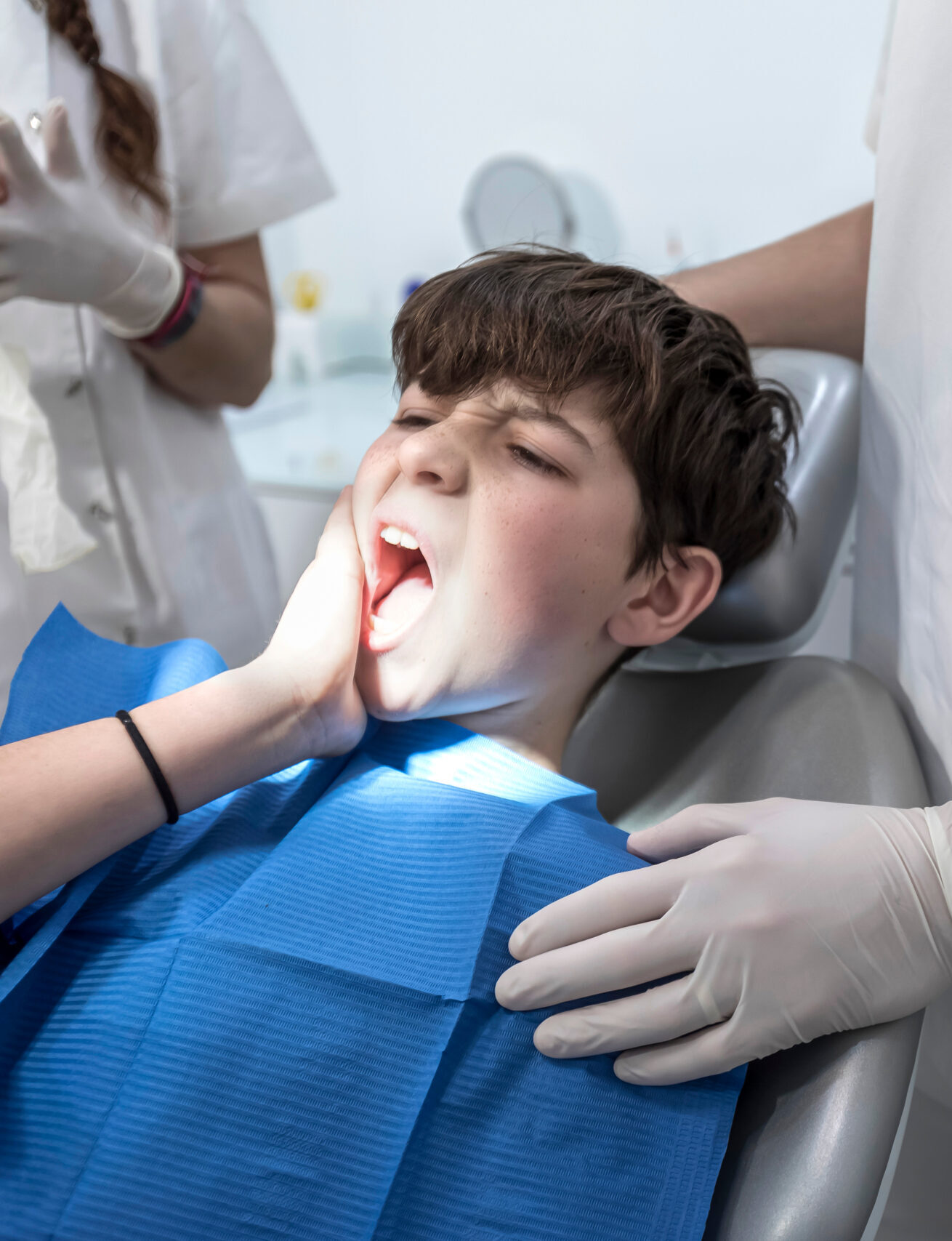 Worried looking boy in the dentist chair in front of the doctor is complaining because of her gum pain and holding her hand on the cheek.