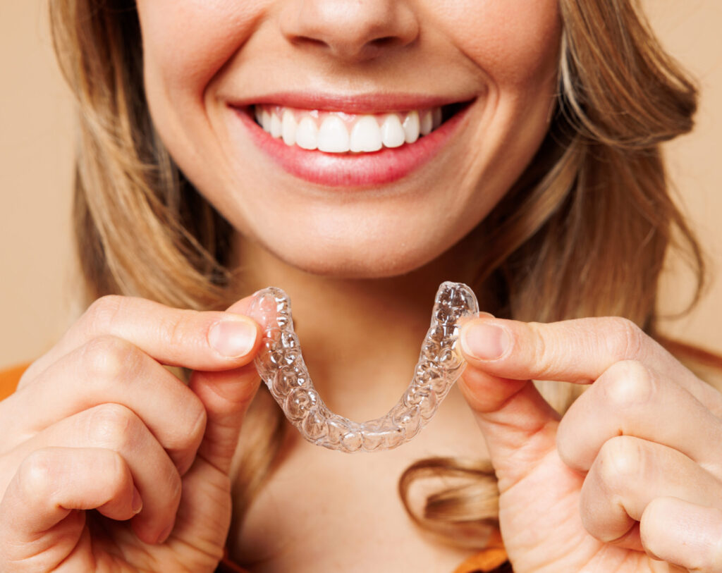 Close up cropped young woman wear orange shirt casual clothes hold in hand invisible transparent aligners, invisalign bracer smile isolated on plain pastel light beige background. Lifestyle concept