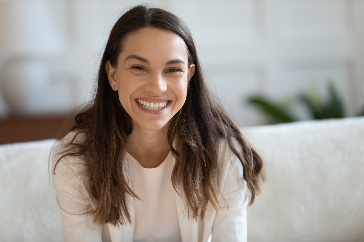 Portrait of happy young mixed race beautiful girl relaxing on cozy sofa. Smiling pretty millennial woman looking at camera, holding video call with friends, laughing at funny joke, profile picture.
